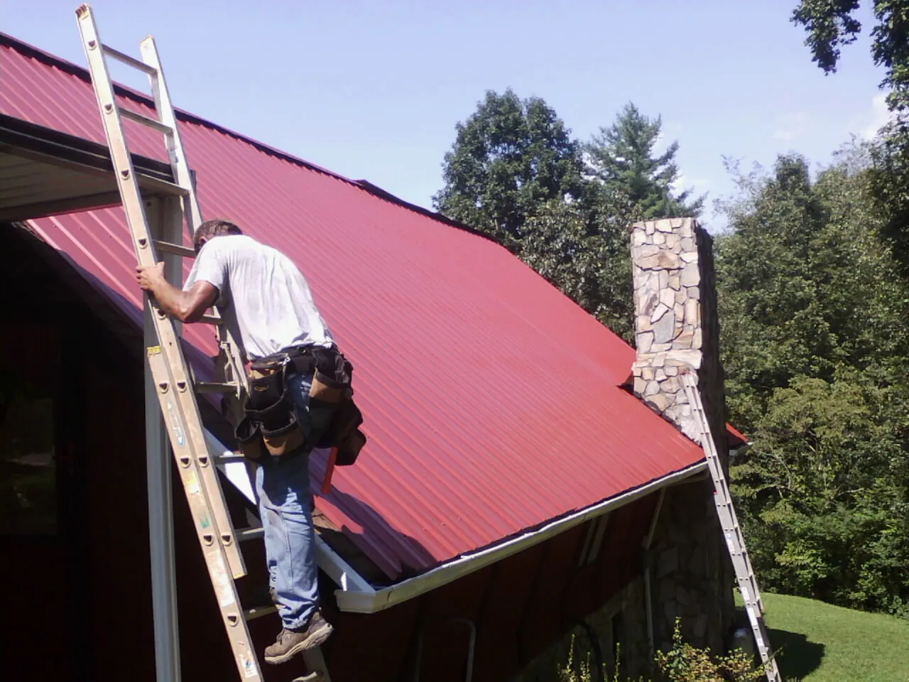 Roofer on a ladder beside a red metal roof and stone chimney — Soffit & Fascia Repair by Roofing Brothers in Chelan
